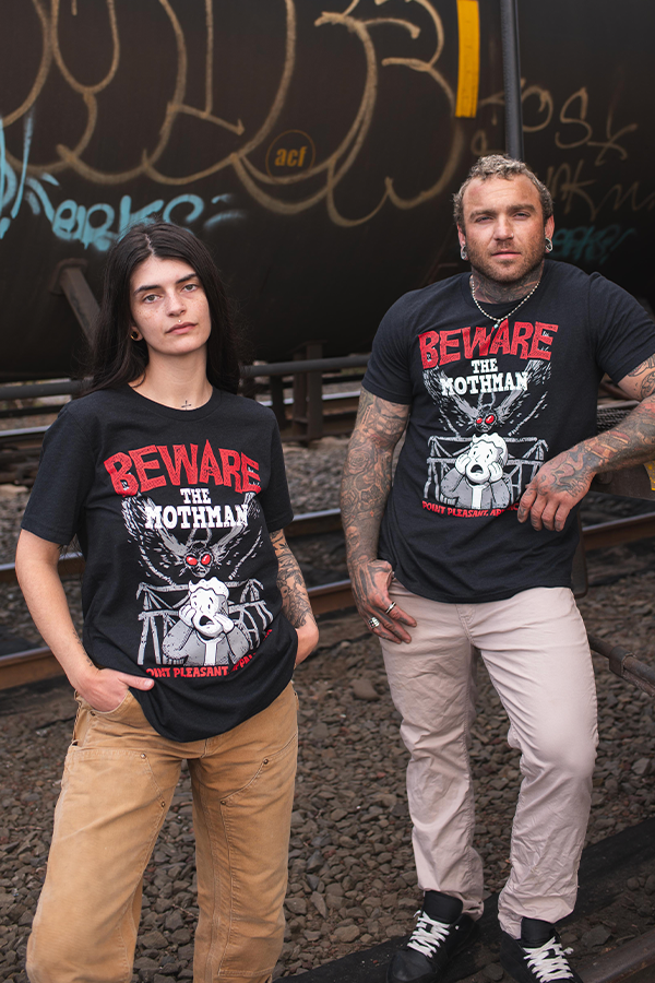 Two people wearing black t-shirts with 'Beware the Mothman' graphic design standing on train tracks with graffiti in the background.