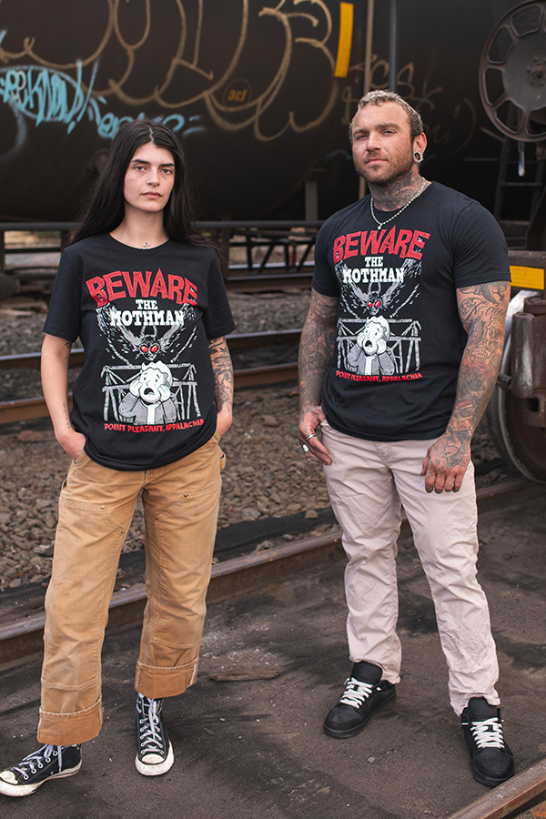 Two people wearing 'Beware the Mothman' t-shirts standing on train tracks with graffiti in the background.