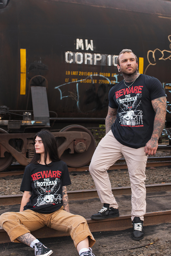 Two people wearing 'Beware the Mothman' t-shirts standing on train tracks with a large train car in the background.